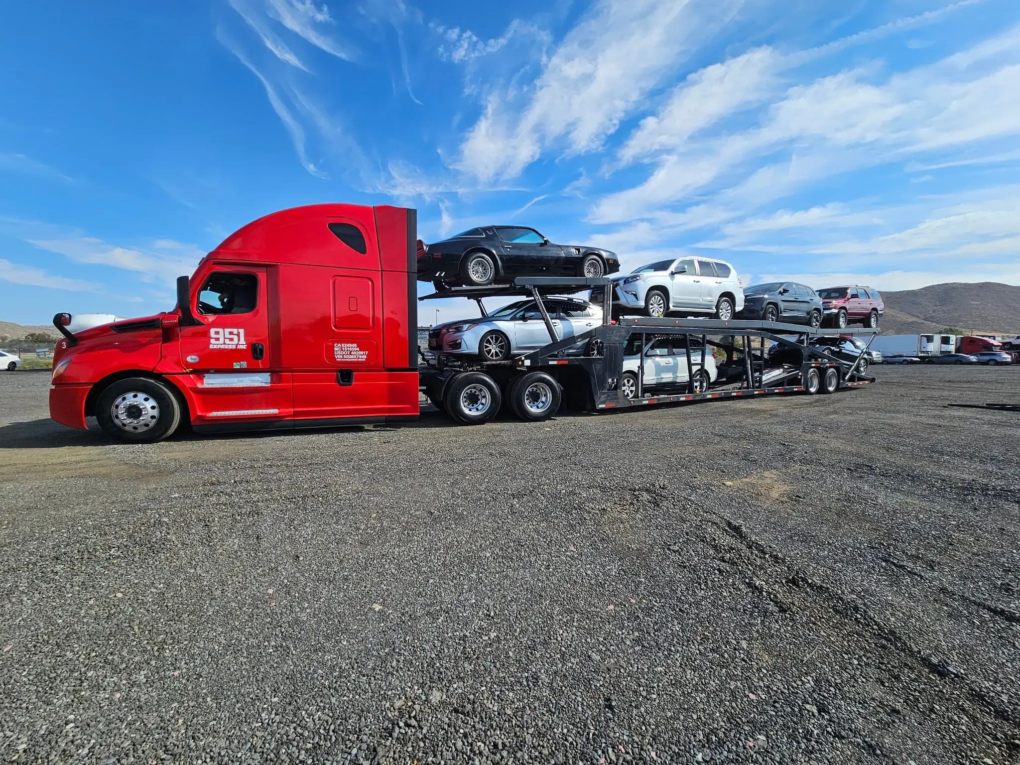 A red 951 Express car hauler loaded with six vehicles, parked on a gravel lot under a blue sky.